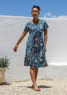 Woman wearing a teal cotton floral dress with v-neck standing against a white wall with blue sky.