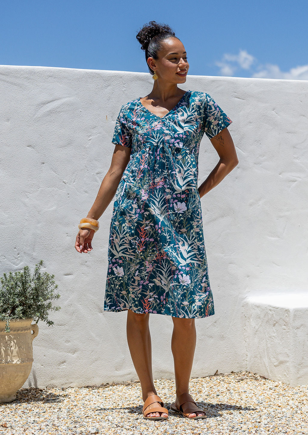 Woman wearing a cotton floral dress with v-neck standing against a white wall with blue sky.