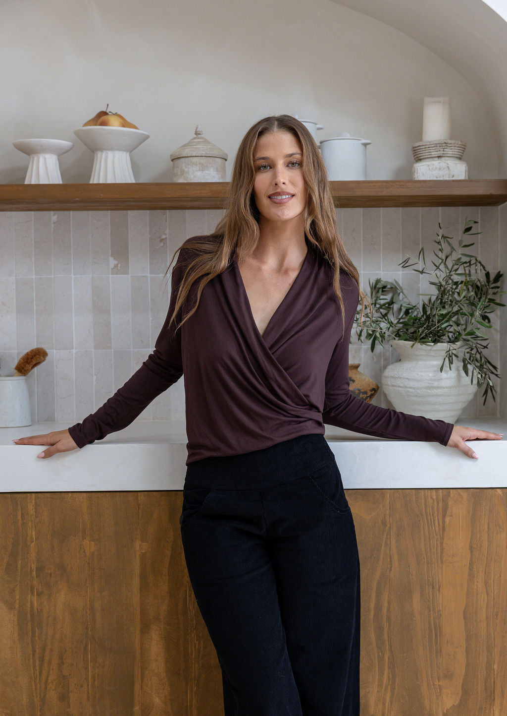 Woman leans on a bench wearing a false wrap front, long sleeve top in dark brown stretch rayon jersey, paired with black pants