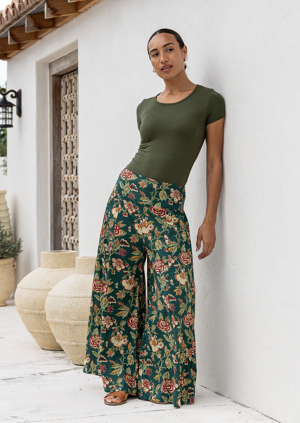 Woman wearing a green t-shirt and wide legged green 100 percent cotton floral pants standing against a white wall.