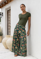 Woman wearing a green t-shirt and wide legged green 100 percent cotton floral pants standing against a white wall.