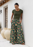 Woman wearing a green top and wide legged green cotton floral pants standing against a white wall.
