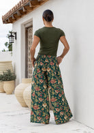 Woman wearing a green top and wide legged green cotton floral pants standing against a white wall back view of hands in pockets