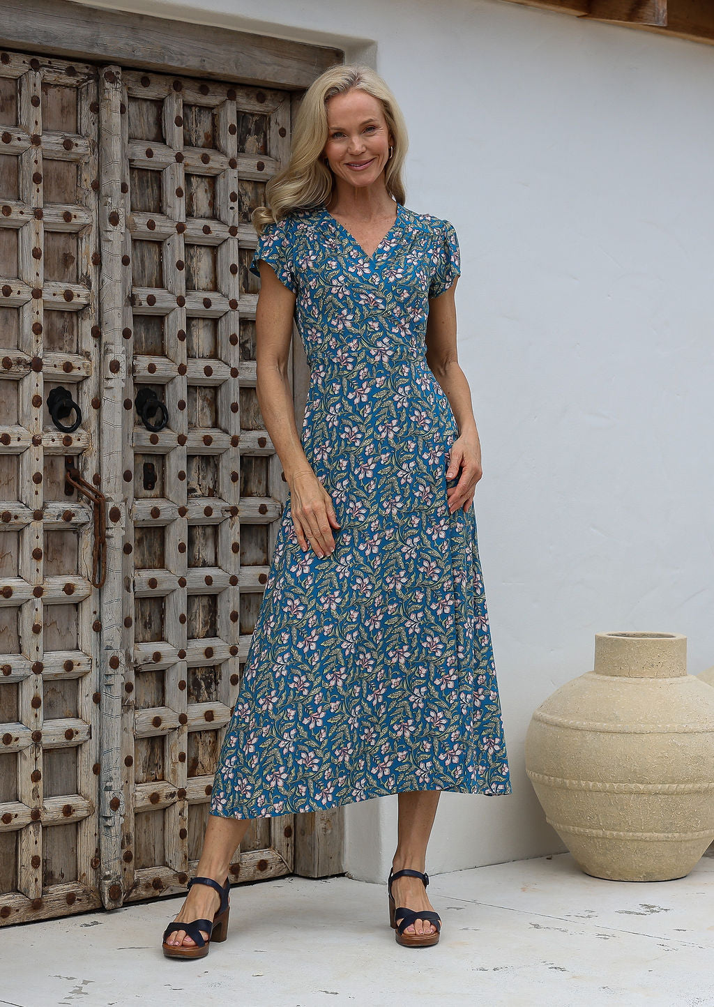 Woman wearing a blue floral print wrap around dress with butterfly sleeves, standing in front of a wooden door