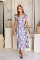 Woman wearing blue based floral print wrap around maxi dress with cap sleeves standing against a white wall