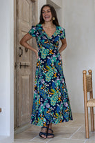Woman wearing a navy blue based floral print wrap around dress with butterfly sleeves, standing in front of a wooden door