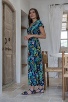 Woman standing sideways wearing a 100% rayon, blue floral wrap around maxi dress with short cap sleeves and long fabric ties to secure the dress