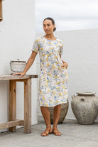 Woman wearing a 1005 cotton floral dress standing next to a wooden table and large vase.