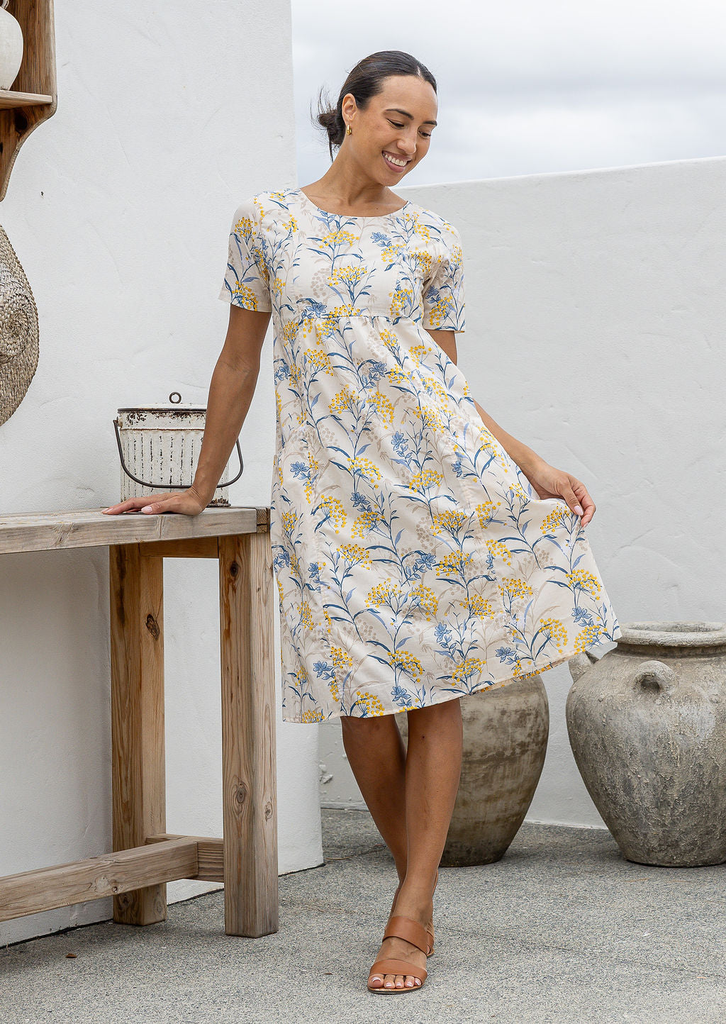 Woman wearing a neutral based floral print cotton dress against a wooden bench and decorative items.