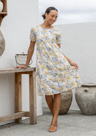 Woman wearing a neutral based floral print cotton dress against a wooden bench and decorative items.
