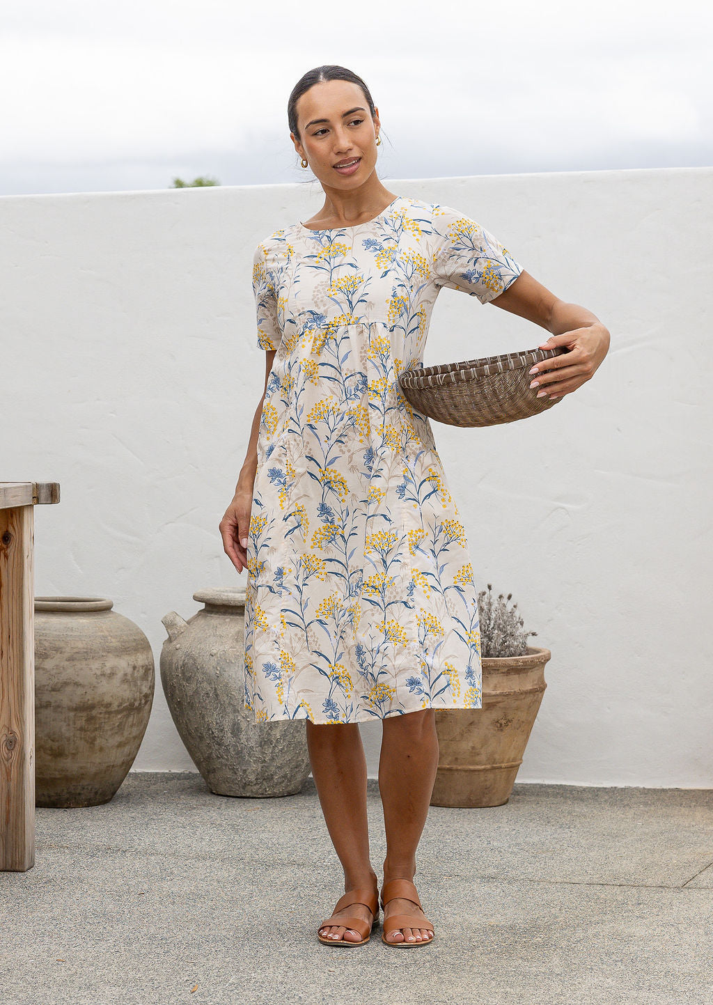 Woman in a cotton floral dress holding a woven basket on a rooftop with potted plants.