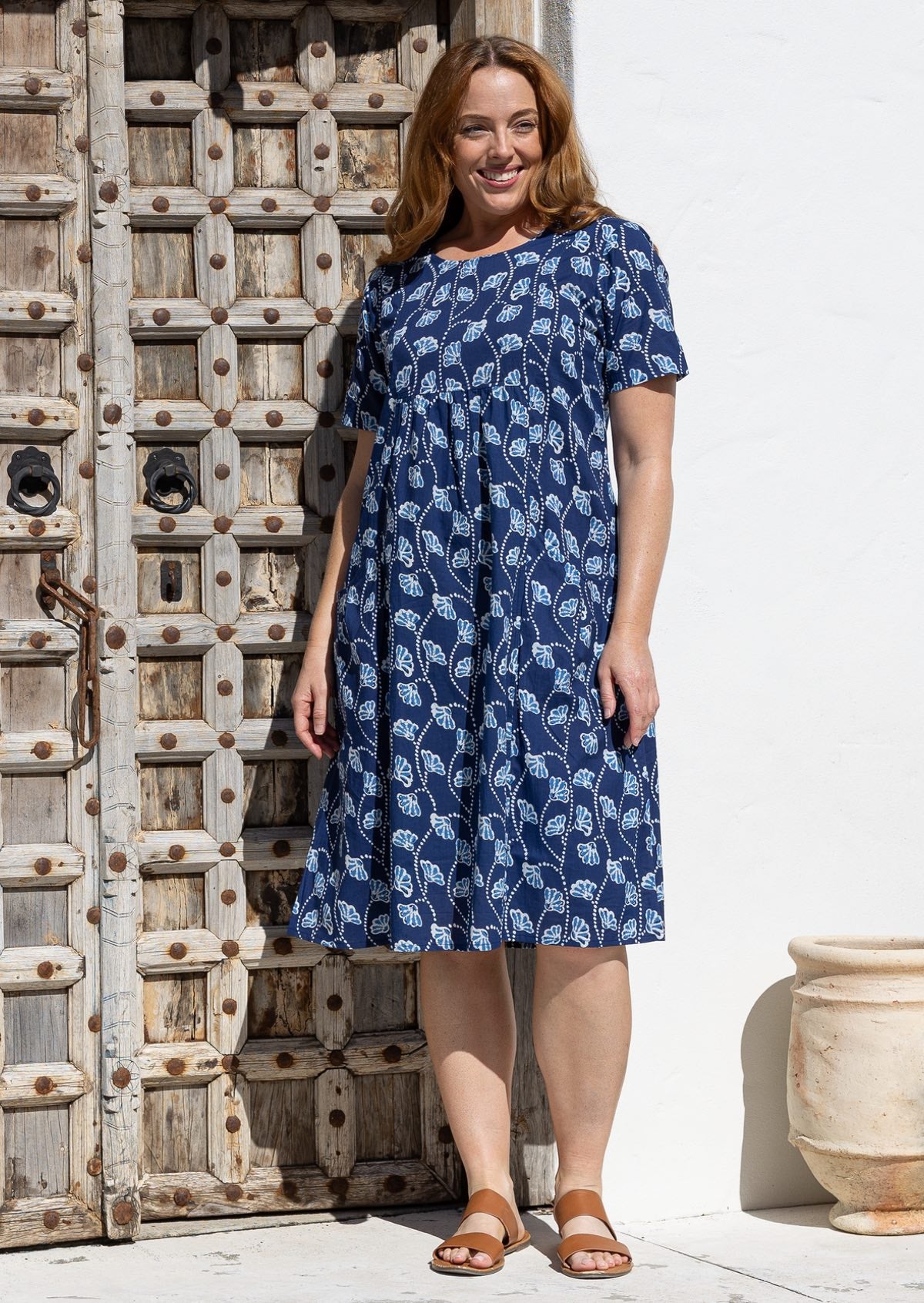 A woman wearing a short-sleeve over the knee-length navy blue cotton dress with a white floral print, standing in front of a rustic wooden door and white wall.