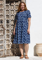 A woman wearing a short-sleeve over the knee-length navy blue cotton dress with a white floral print, standing in front of a rustic wooden door and white wall.