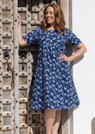 A woman wearing a navy blue short-sleeve cotton dress with a white floral print, standing in front of a rustic wooden door and white wall.