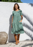 Woman wearing a light sage green cotton dress with a white wall and pot in the background
