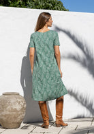 Woman stands backwards wearing a 100% cotton dress in sage green based floral print with short sleeves and brown boots