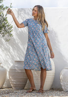 A woman wearing a blue short-sleeve cotton dress floral print, standing outdoors in front of a white wall.