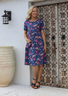 A woman stands with one hand in pocket wearing a 100% cotton, blue based floral print dress with a loose fit style, short sleeves and a round neckline.