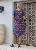 Woman in a blue floral cotton dress with short sleeves, standing in front of a wooden door.