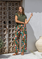 Woman standing wears 100% cotton wide leg floral print pants in olive green based with elastic waist, pockets and pleated ankles