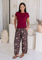 Woman wearing a maroon sleeveless top and black based floral print wide leg cotton pants with pleated ankles, elastic waist and pockets