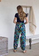 Woman stands backwards wearing a 100% cotton wide leg pants in navy based multicolour floral print with high waist elastic.