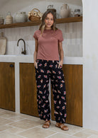 Woman standing in a kitchen wears 100% cotton wide leg floral print pants in black with elastic waist, pockets and pleated ankles