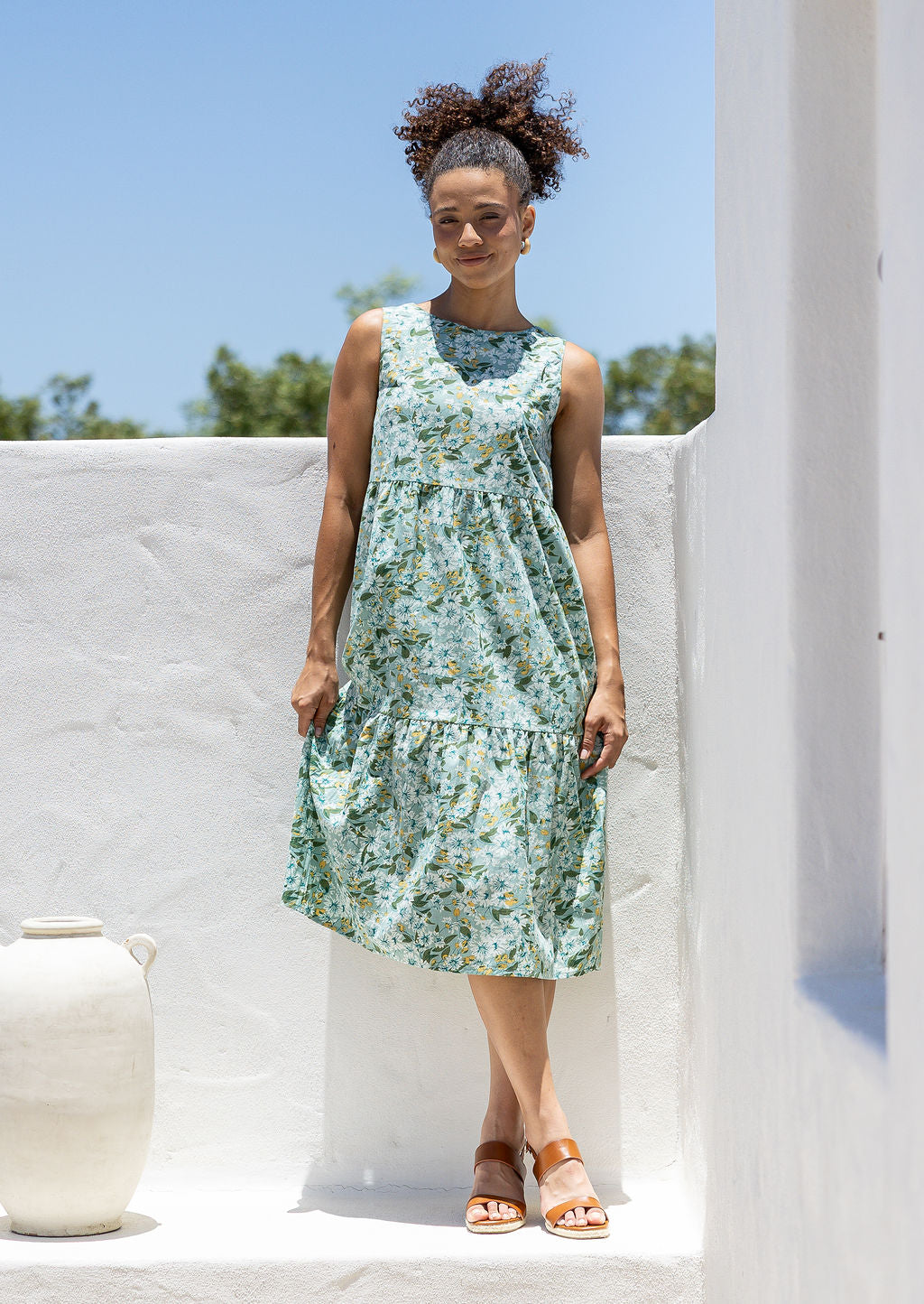 Woman standing against the white wall is wearing tiered midi length dress with relaxed fit, empire waist and pockets gorgeous sage green based floral print
