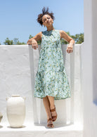 Woman in a cotton floral dress standing against a white wall with blue sky and greenery.