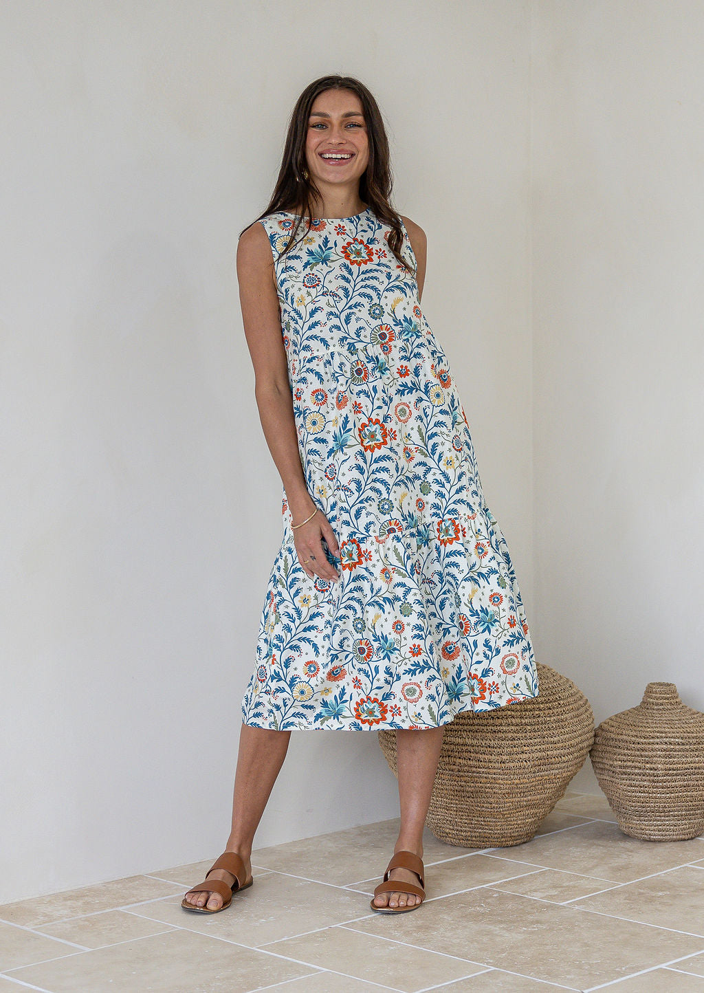 Model poses in sleeveless, white based floral print cotton midi dress with tiered design, pockets and high round neckline.