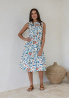 Woman standing against the white wall is wearing tiered midi length dress with relaxed fit, empire waist and pockets gorgeous white based floral print