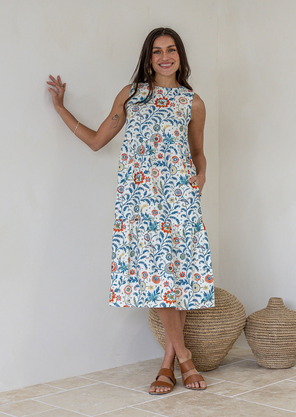 A woman wearing a sleeveless midi dress with a white based floral print standing against a white wall