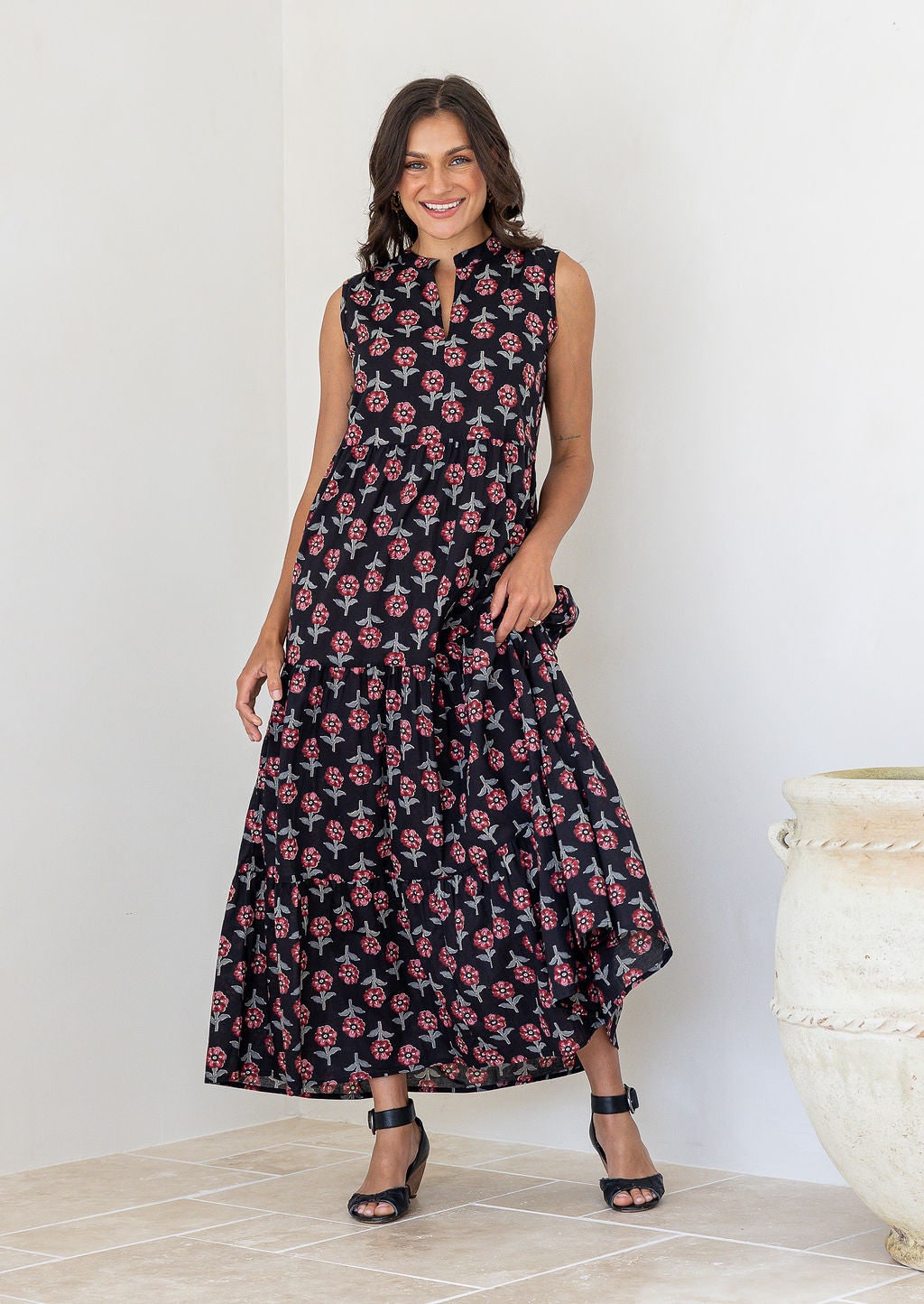 woman with dark hair wearing black floral tiered cotton maxi dress, sleeveless with notch neckline, worn by model with black heeled sandals in white room