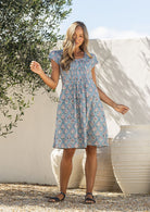 Woman poses in a 100% cotton blue based floral dress with round neckline and cap sleeves