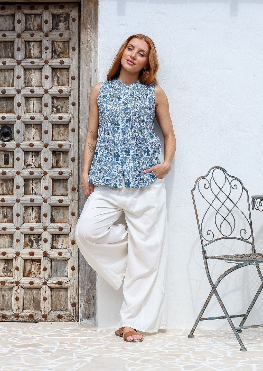 Woman posing wearing a cotton sleeveless top in white based blue floral print with small decorative pleats in the front.