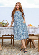 Woman stands wearing a 100% cotton dress in white based blue floral print with short sleeves and one hand in pocket