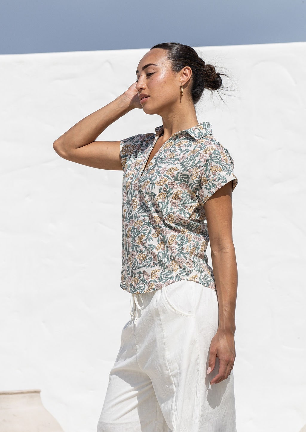 Woman stands sideways wearing a white based relaxed fit, shirt style floral cotton top. 