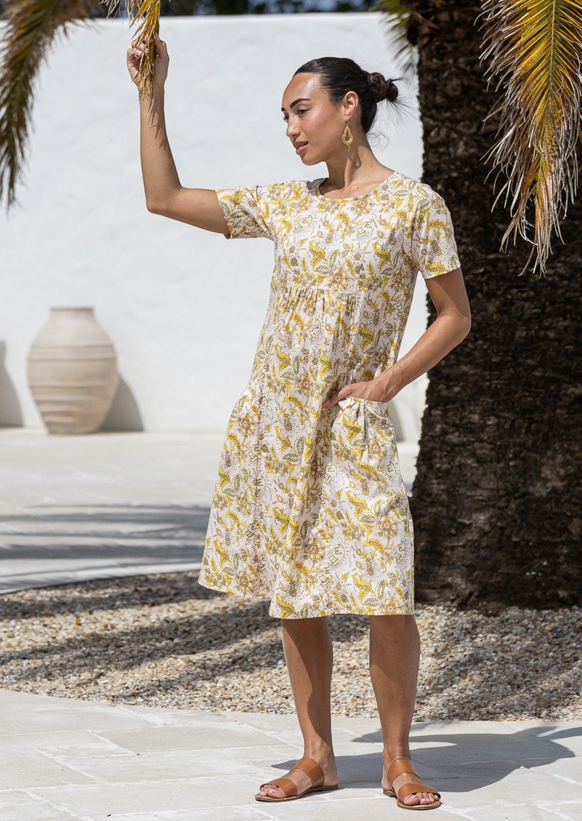 A woman wearing a cream short-sleeve cotton dress floral print with one hand in pocket, standing outdoors. 