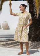 A woman wearing a cream short-sleeve cotton dress floral print with one hand in pocket, standing outdoors. 