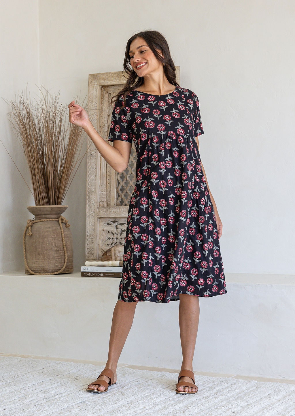 A woman wearing a black short-sleeve cotton dress floral print, standing in front of a rustic wooden door and white wall.