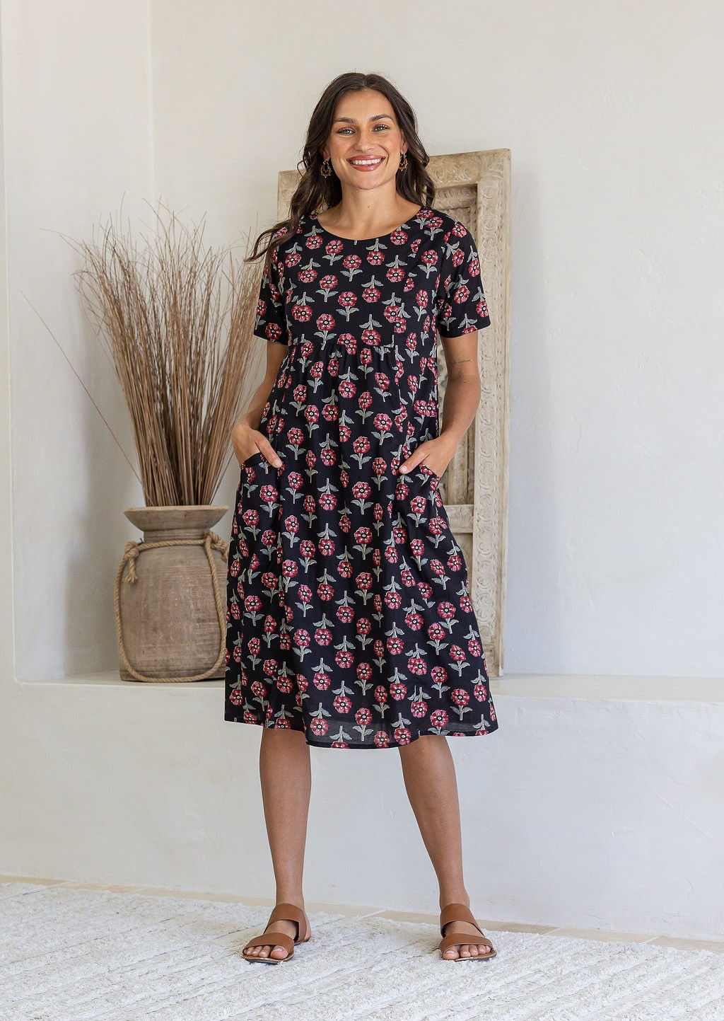 Woman stands wearing a 100% cotton dress in black based floral print with short sleeves and both hand in pocket