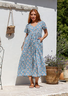 Woman standing in a 100% cotton floral print dress with a V-neckline, short sleeves, pockets, and buttons through to the knee.