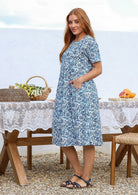 Woman stands sideways wearing a 100% cotton dress in white based blue floral print with short sleeves and one hand in pocket