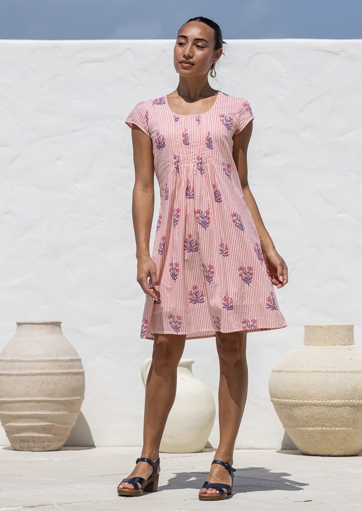 Woman poses in a 100% cotton pink striped white based floral dress with round neckline and cap sleeves