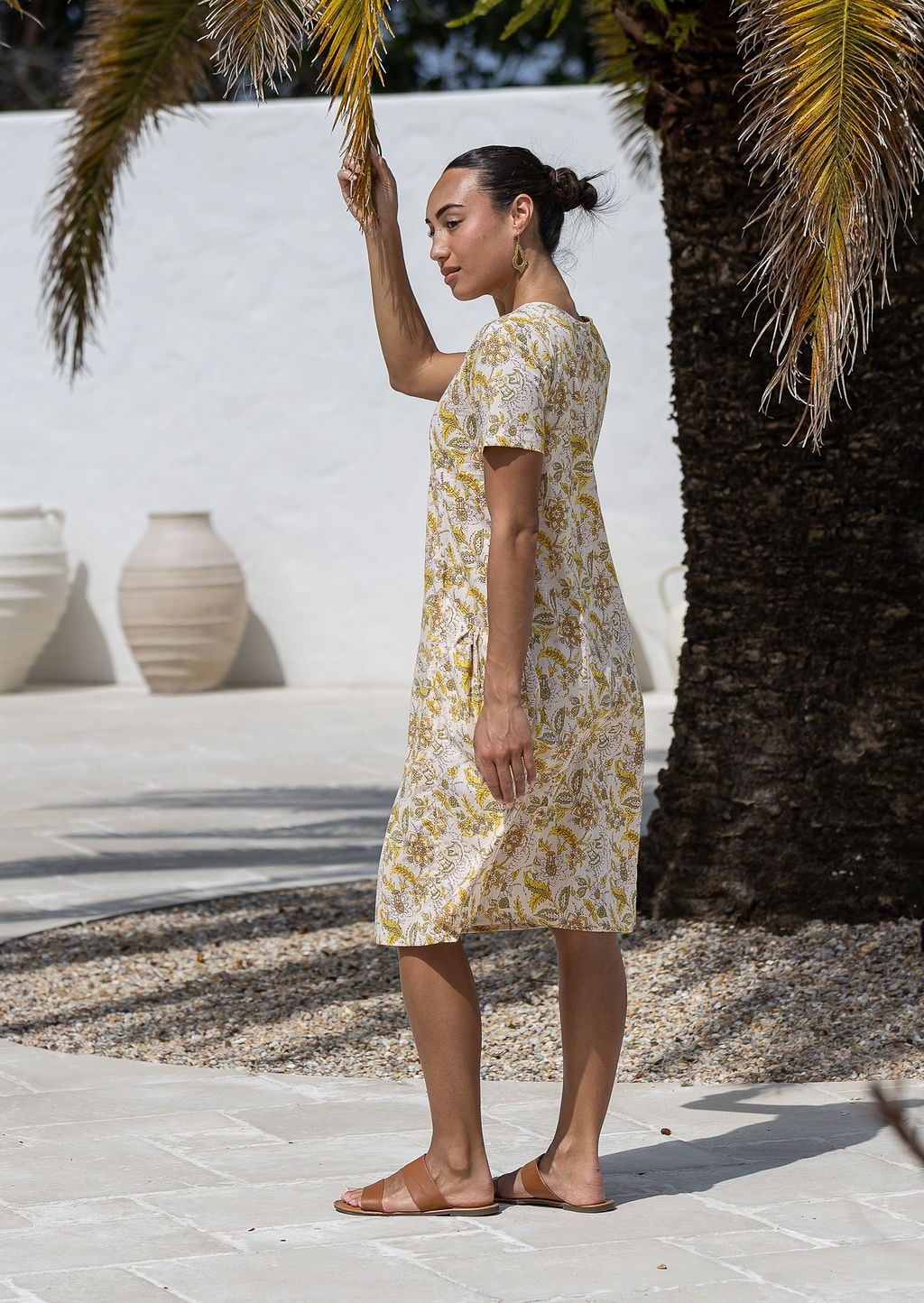 Woman stands sideways wearing a 100% cotton dress in cream based floral print with short sleeves. 