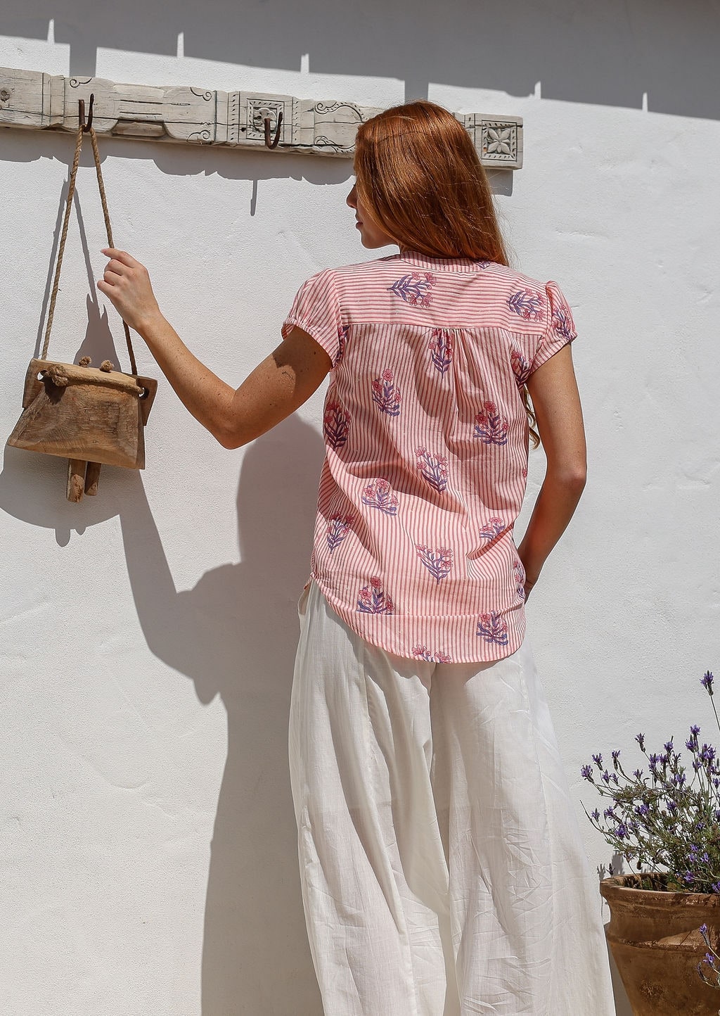 Woman stands backwards wearing a 100% cotton pink striped floral short sleeve top and plain white pants.