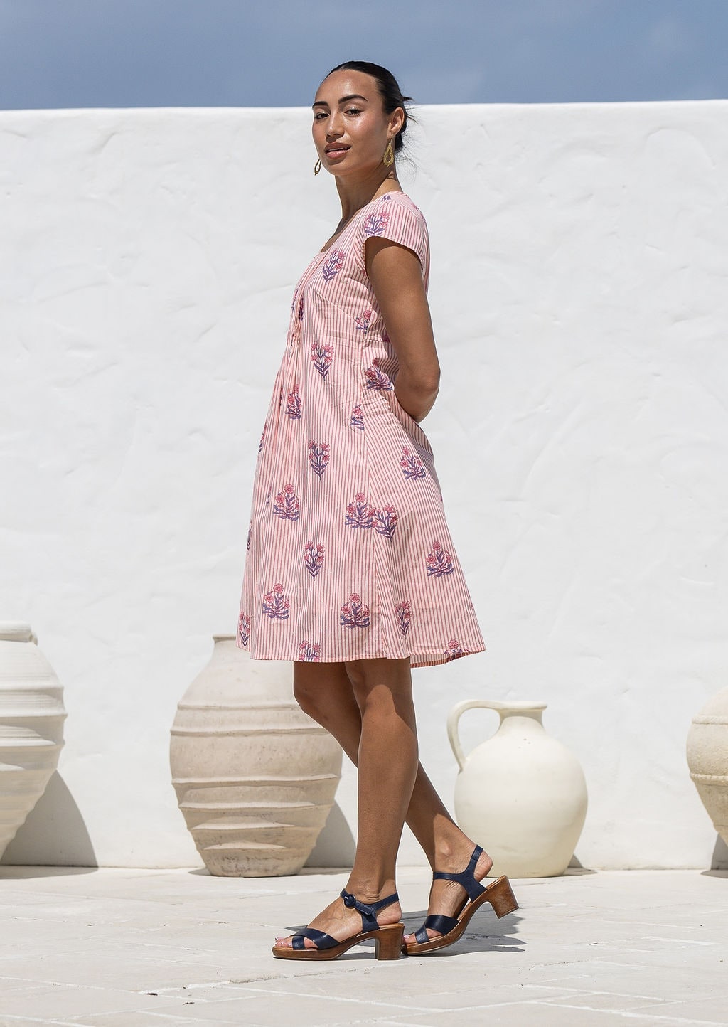 Woman stands sideways wearing a 100% cotton pink striped white based floral short-sleeve dress with round neckline.