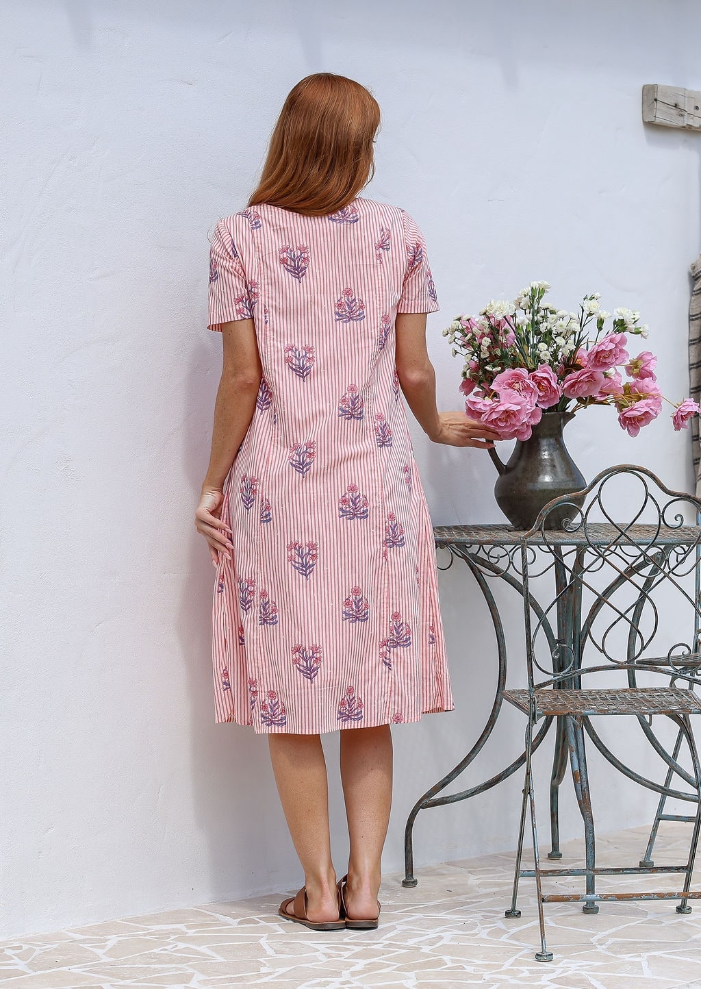 Woman stands backwards wearing a 100% cotton dress in pink striped white based floral print with short sleeves paired with brown sandals. 
