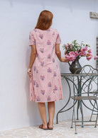 Woman stands backwards wearing a 100% cotton dress in pink striped white based floral print with short sleeves paired with brown sandals. 