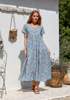 Woman standing in a 100% cotton floral print dress with a V-neckline, short sleeves, pockets, and buttons through to the knee.